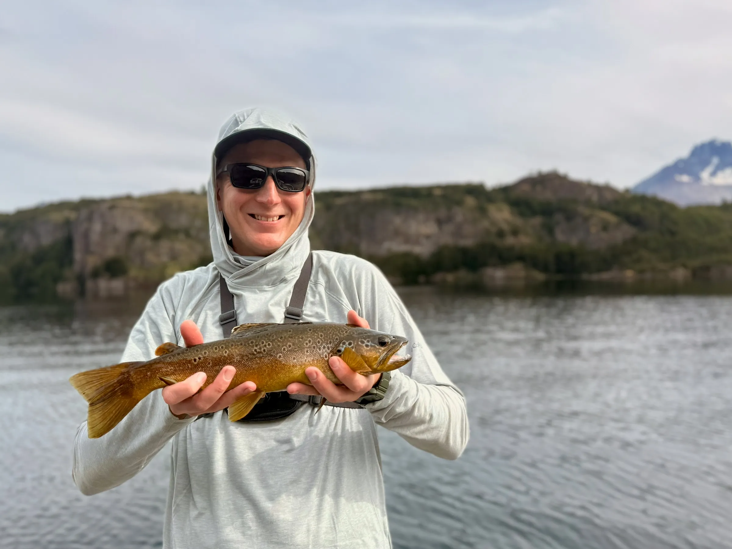 A grinning angler holding a golden brown trout, lake and mountains behind