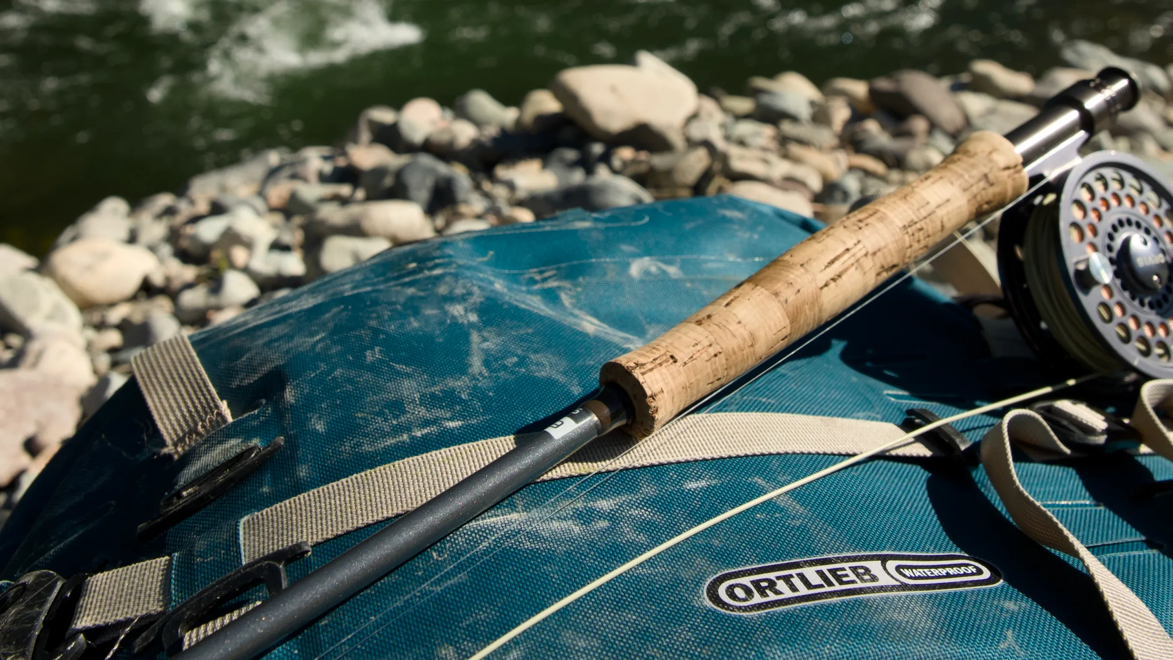 Fly rod and reel resting on a well-worn Ortlieb Atrack by the river
